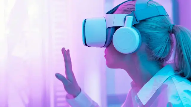 Woman wearing a virtual reality headset and headphones raising one hand in a setting lit in pink and violet