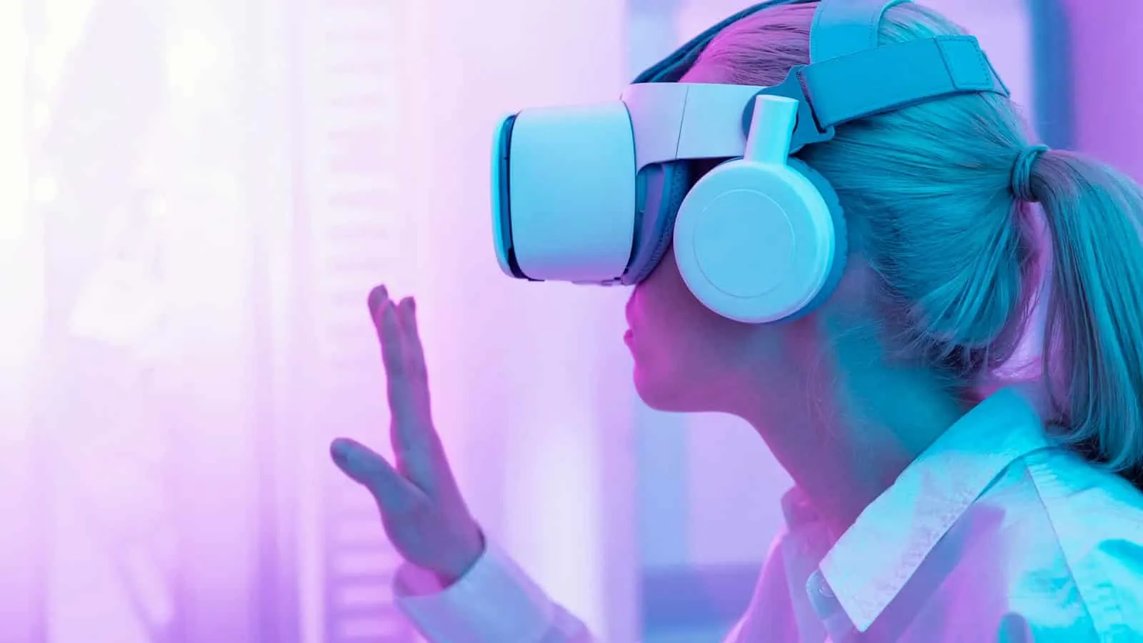 Woman wearing a virtual reality headset and headphones raising one hand in a setting lit in pink and violet