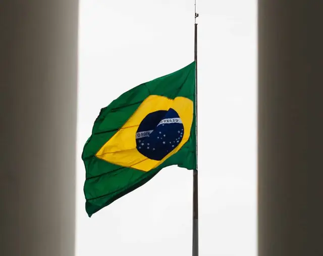 Brazilian flag flying at half-mast against a white sky, framed by two dark walls