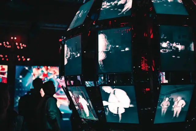 Rotating cube of screens showing black-and-white figures, with silhouettes of people in a dark room, like a surveillance image