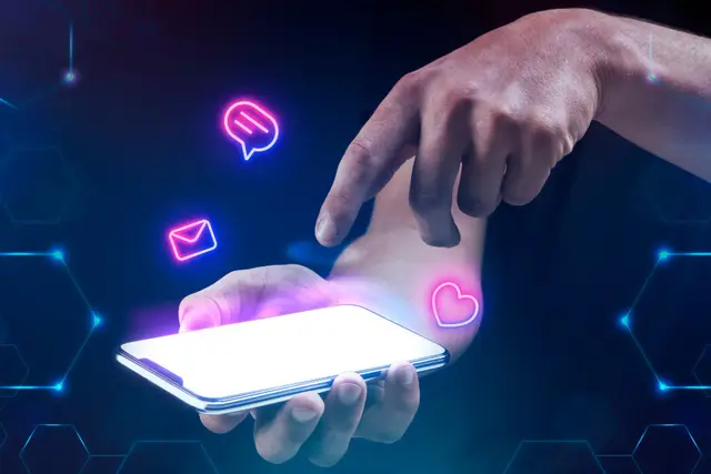Hand touching a phone screen with message, mail and heart icons in pink neon floating over a dark blue background.