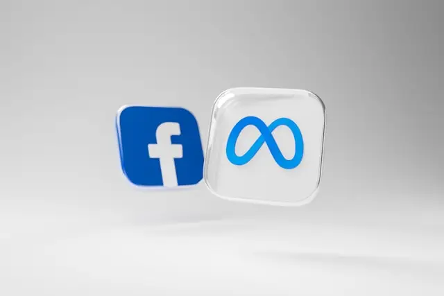 Two floating 3D icons on a light gray background: the blue Facebook logo and the Meta infinity symbol on a translucent cube.