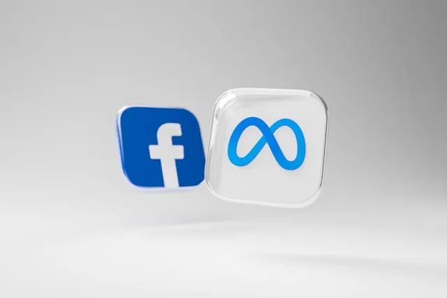 Two floating 3D icons on a light gray background: the blue Facebook logo and the Meta infinity symbol on a translucent cube.