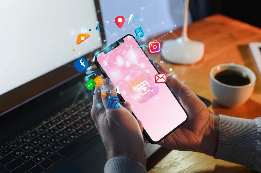 Desk scene with a phone showing a pink screen and app icons floating above it, with a laptop and a coffee cup behind.