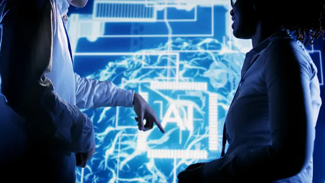 Two people stand before a blue screen with the letters AI over a glowing brain, one of them pointing at its center.