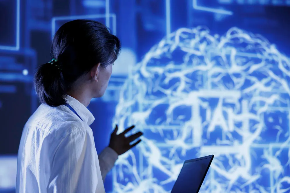Woman in profile raising a hand before a large glowing blue brain, with a laptop in the foreground and the letters AI in the image.