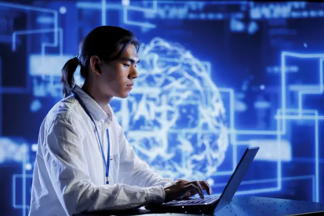 Young person in profile typing on a laptop in front of a large glowing brain made of blue lines on a technological background.
