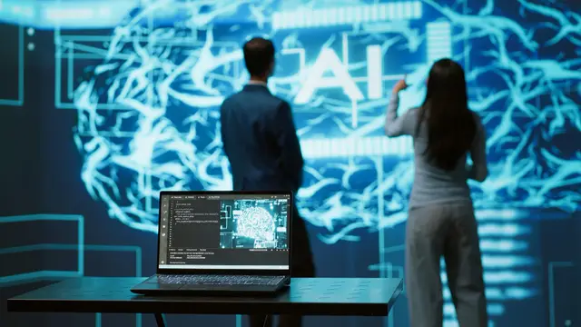 A man in a suit and a woman seen from behind stand before a large glowing brain with the letters AI, with a laptop in the foreground.