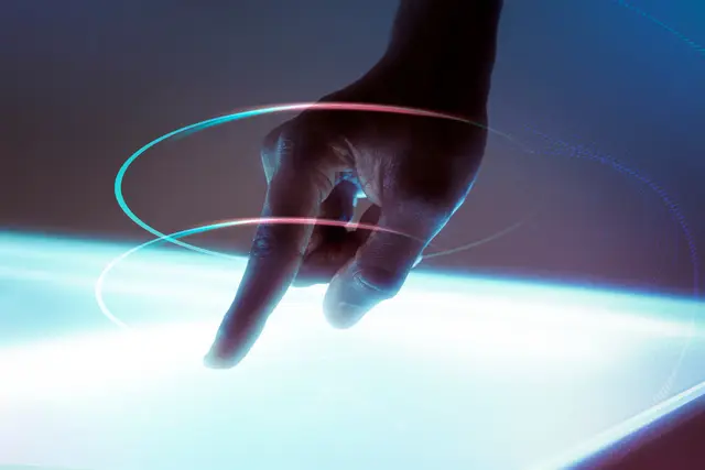 A hand with an extended finger touches a glowing surface, ringed by two loops of pink and turquoise light.