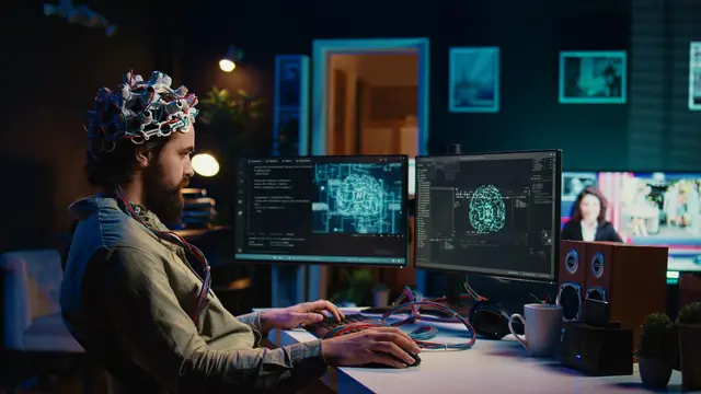Man wearing an electrode cap sits before two monitors displaying brain images, in a dimly lit office.