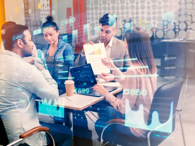 Four professionals work with laptops and papers at a table, seen through glass over which glowing charts and figures float.