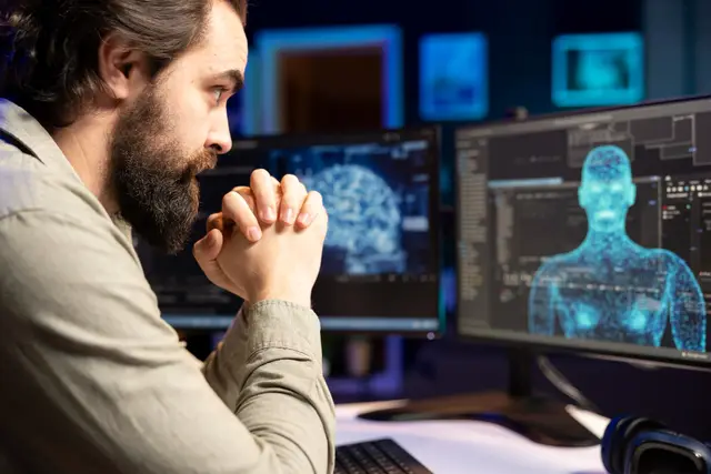 Bearded man in profile with clasped hands in front of two monitors showing a digital brain and a 3D human figure