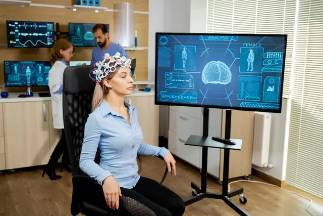Woman wearing an electrode cap to record brain activity, seated before a screen showing a brain and data, while two researchers work in the background.