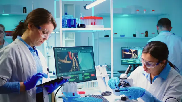 Two scientists in lab coats and gloves work in a blue-lit laboratory with a tablet, a microscope and a monitor