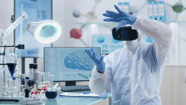 Person in a protective suit and virtual reality headset raising gloved hands in front of a monitor showing a brain