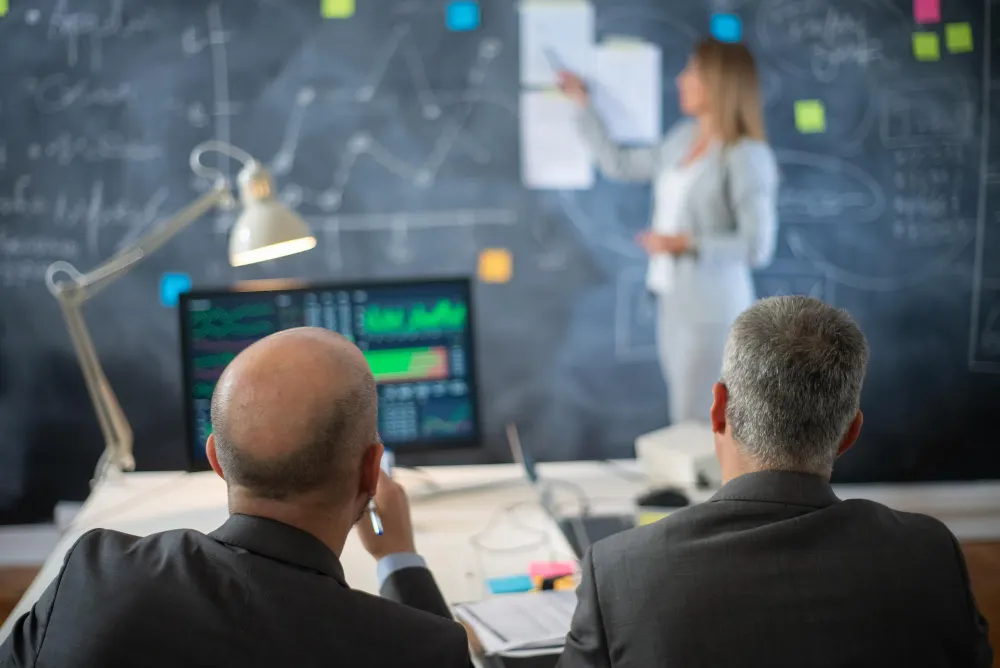 Two men seen from behind watch a woman presenting at a chalkboard covered in notes, with a monitor of charts in the foreground