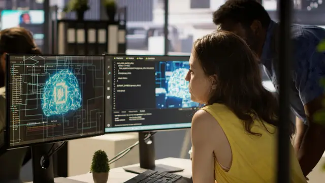 Woman in a yellow top and a man behind her look at two monitors showing digital brains and code in an office