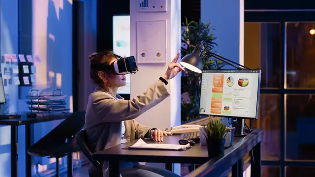 Woman wearing a virtual reality headset raising her hand at a desk with a monitor of charts, in an office at night