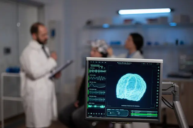 Scientist in a white coat and a patient wearing an electrode cap in the background, with a monitor in the foreground showing a 3D brain