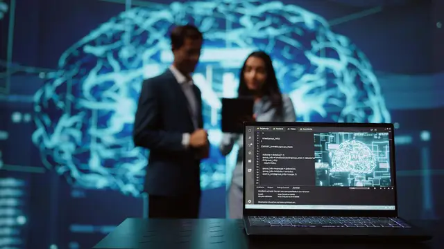 A man and a woman standing out of focus in front of a glowing projection of a brain, with a laptop showing code in the foreground