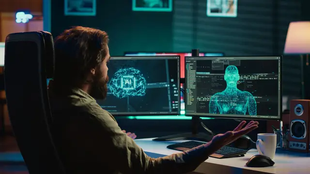 Bearded man at a desk with open hands in front of two monitors showing a digital brain and a 3D human figure