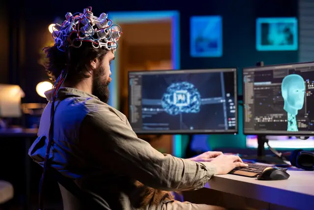 Bearded man wearing an electrode cap in front of two monitors showing a brain and a digital human figure