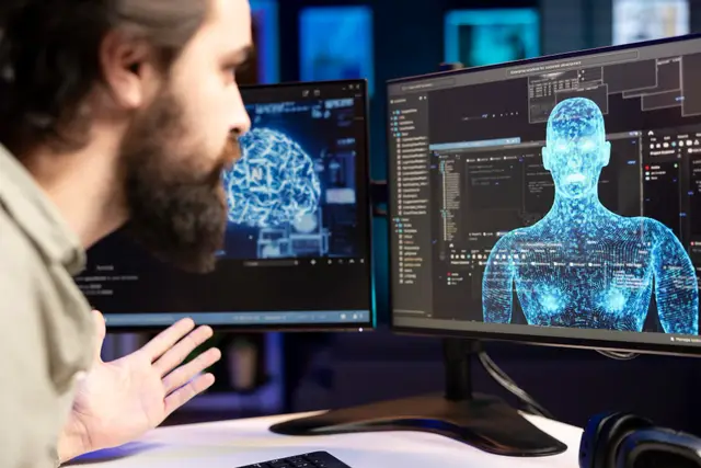 Bearded man in profile gesturing in front of two monitors, one showing a brain and the other a 3D human figure