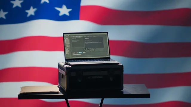 Open laptop on a black case on a small table in front of a large American flag.