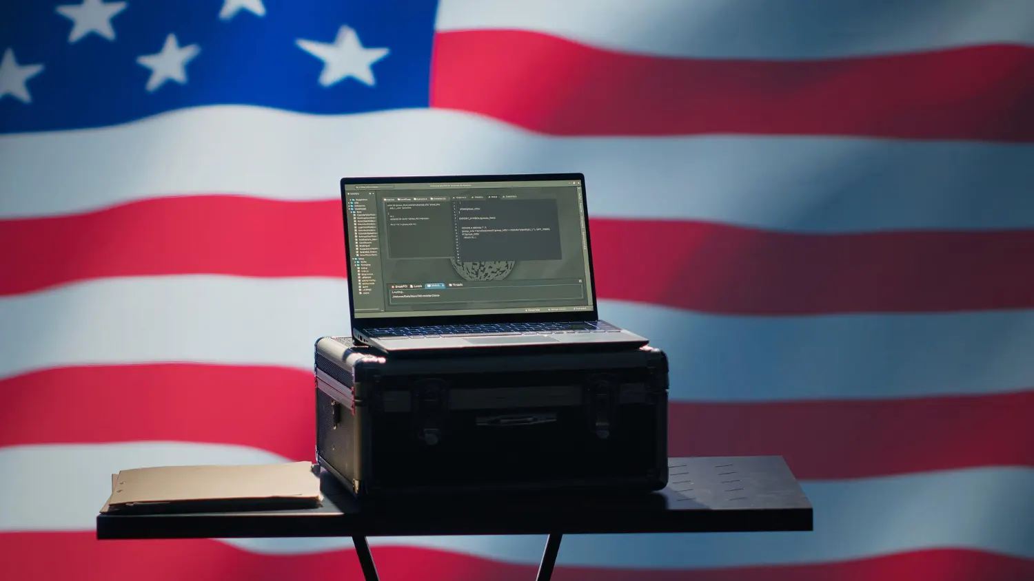 Open laptop on a black case on a small table in front of a large American flag.