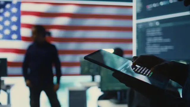 Silhouette of a man in front of a U.S. flag and screens full of code, with a tablet held in the foreground