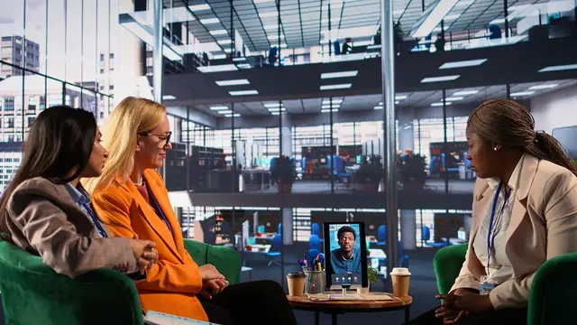 Three women talk in a modern office beside a table with a tablet showing a video call
