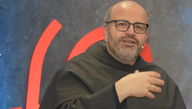 Close-up of a bespectacled Franciscan friar in a dark habit speaking and gesturing in front of a background with red shapes