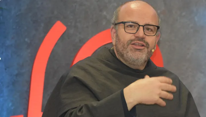 Close-up of a bespectacled Franciscan friar in a dark habit speaking and gesturing in front of a background with red shapes