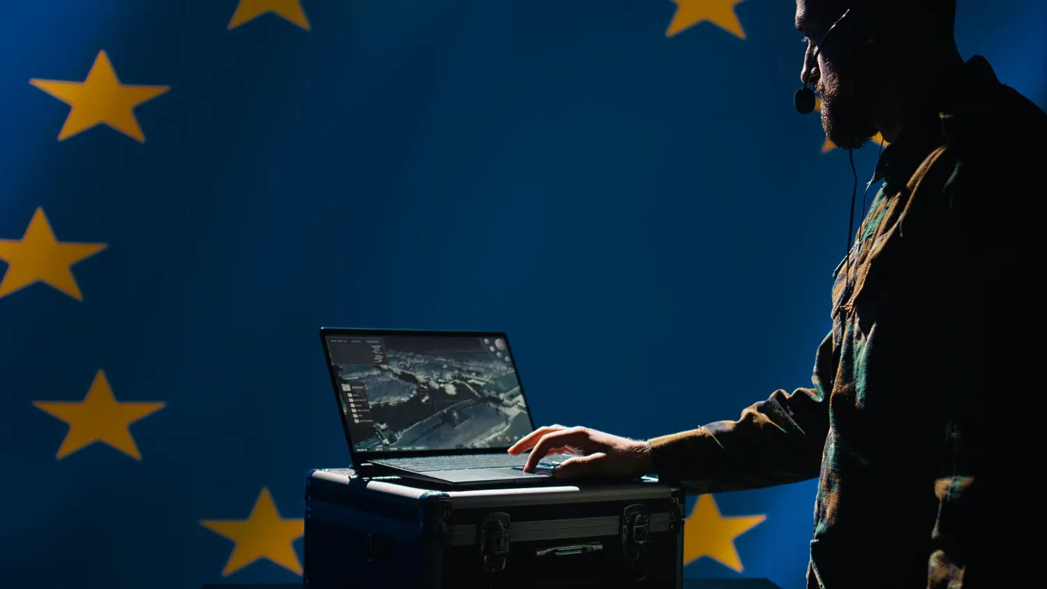 Man wearing a headset types on a laptop in front of a blue European Union flag with yellow stars