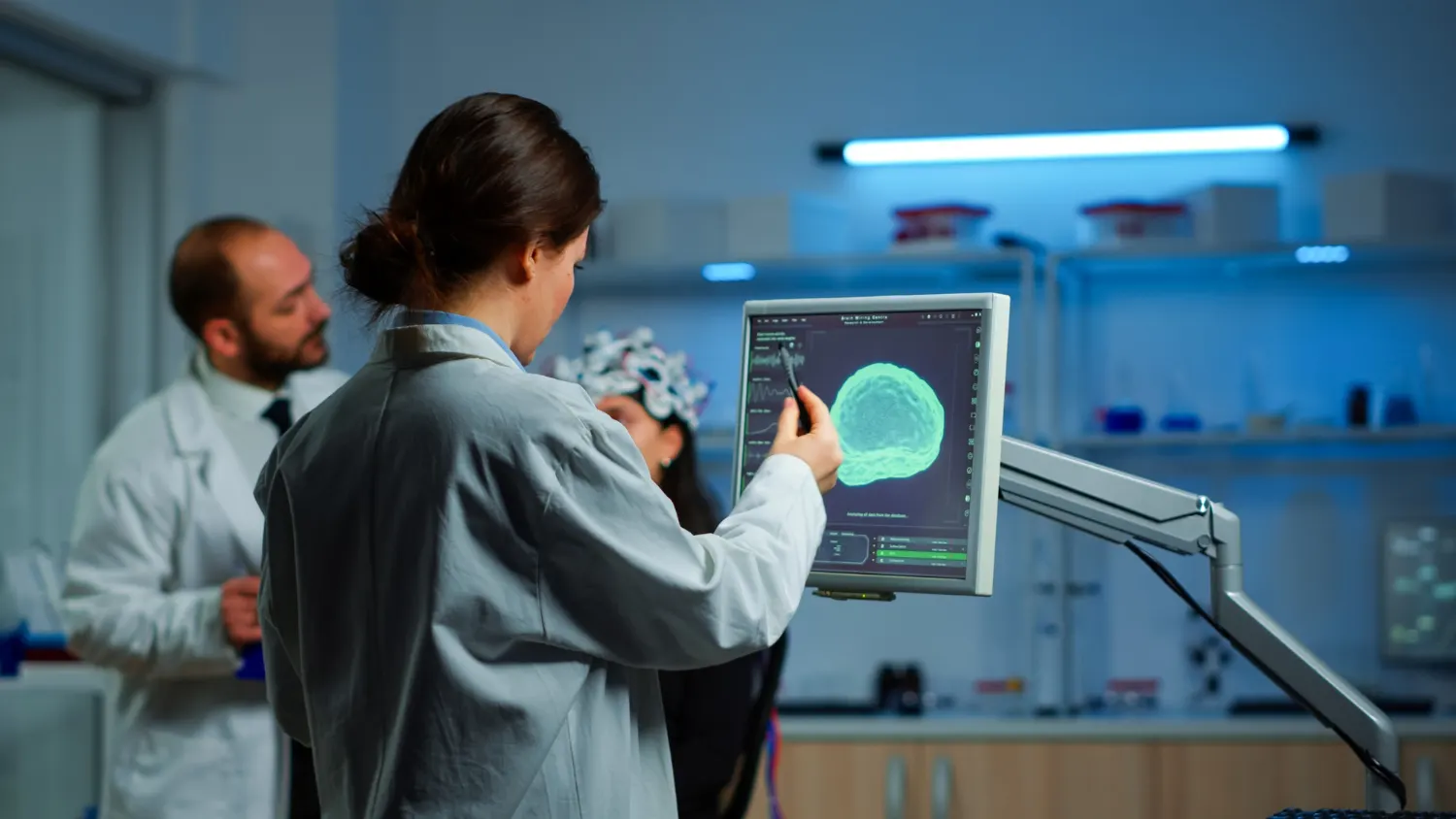 A scientist points at a screen showing a green brain image while a colleague wearing an electrode cap looks on in a laboratory
