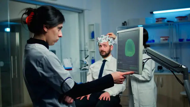 Researchers in a laboratory point at a screen showing a brain scan while a colleague wears an electrode cap