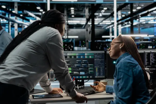 Two women working in front of data monitors in a server room, one leaning over the desk