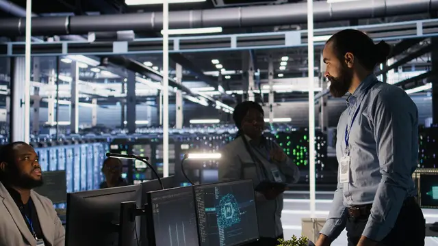 A man talks with colleagues in front of monitors in a bright server room
