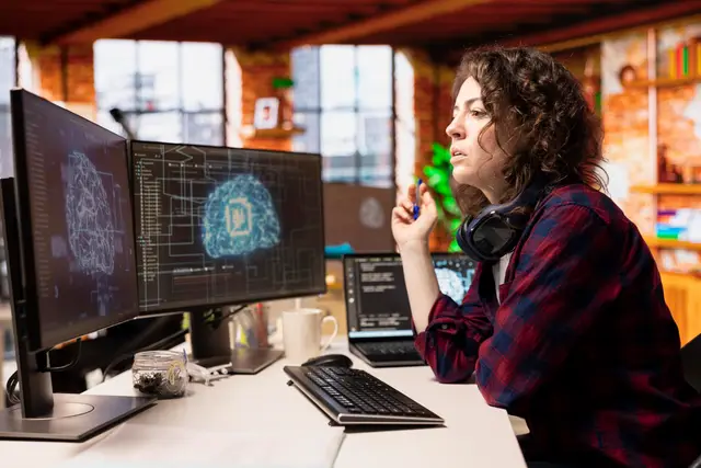 Thoughtful woman in front of monitors showing a digital brain and code, in an office with exposed brick