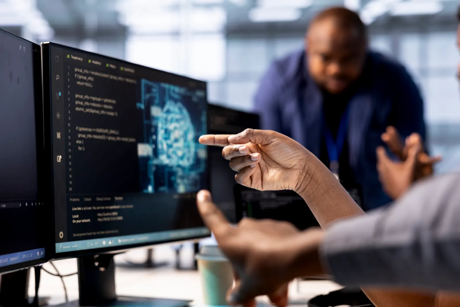 Several people point at a monitor showing a digital brain and code while they discuss in a work room