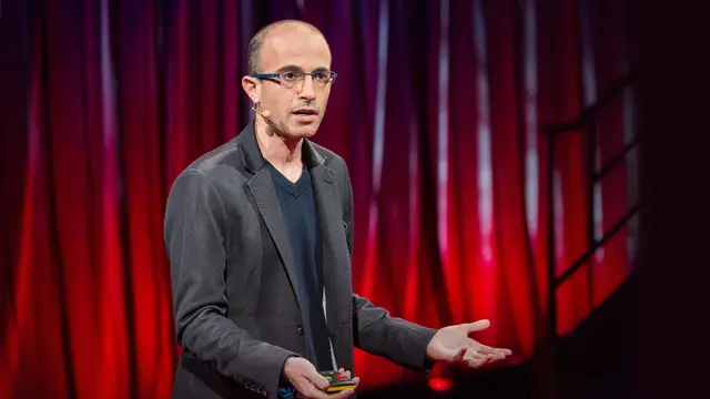 Man in glasses and a gray jacket speaks with a headset microphone and open hands in front of a red curtain on a stage
