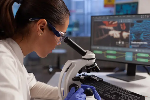 Researcher in safety glasses looks through a microscope beside a monitor showing cell images in a laboratory