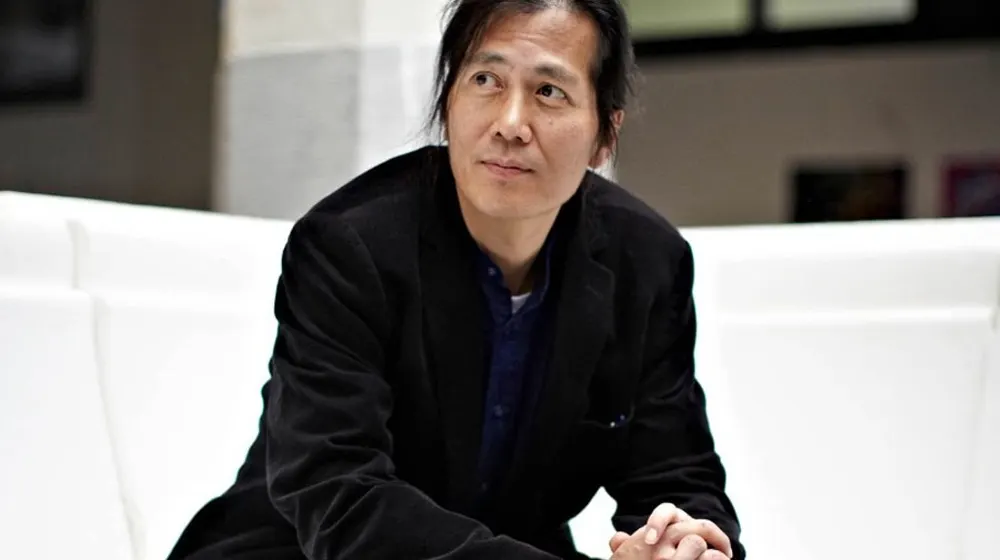 Man with long dark hair and a black jacket, seated on a white sofa with his hands clasped, looking off to one side