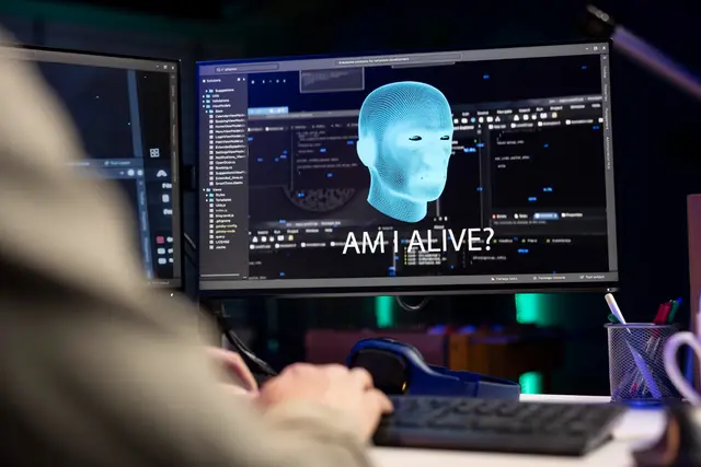 Monitor showing a blue digital head and the text “Am I alive?” in front of a person typing, with more code screens nearby