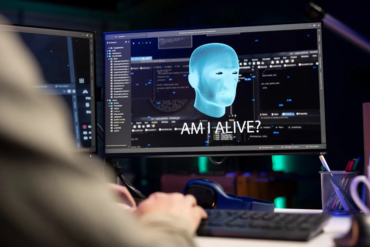 Monitor showing a blue digital head and the text “Am I alive?” in front of a person typing, with more code screens nearby