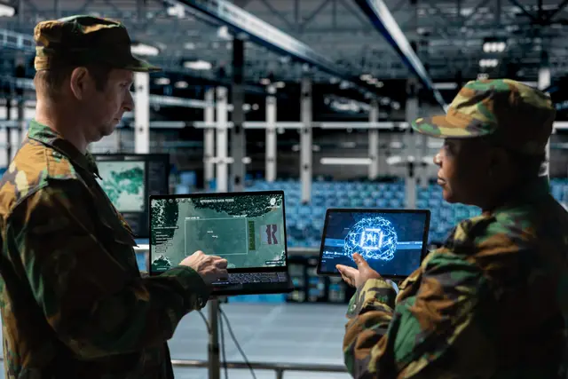 Two soldiers in camouflage uniforms review maps and a digital brain graphic on laptops inside a large hangar