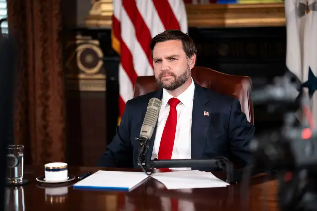 Bearded man in a dark suit and red tie seated at a microphone in an office, with American flags in the background