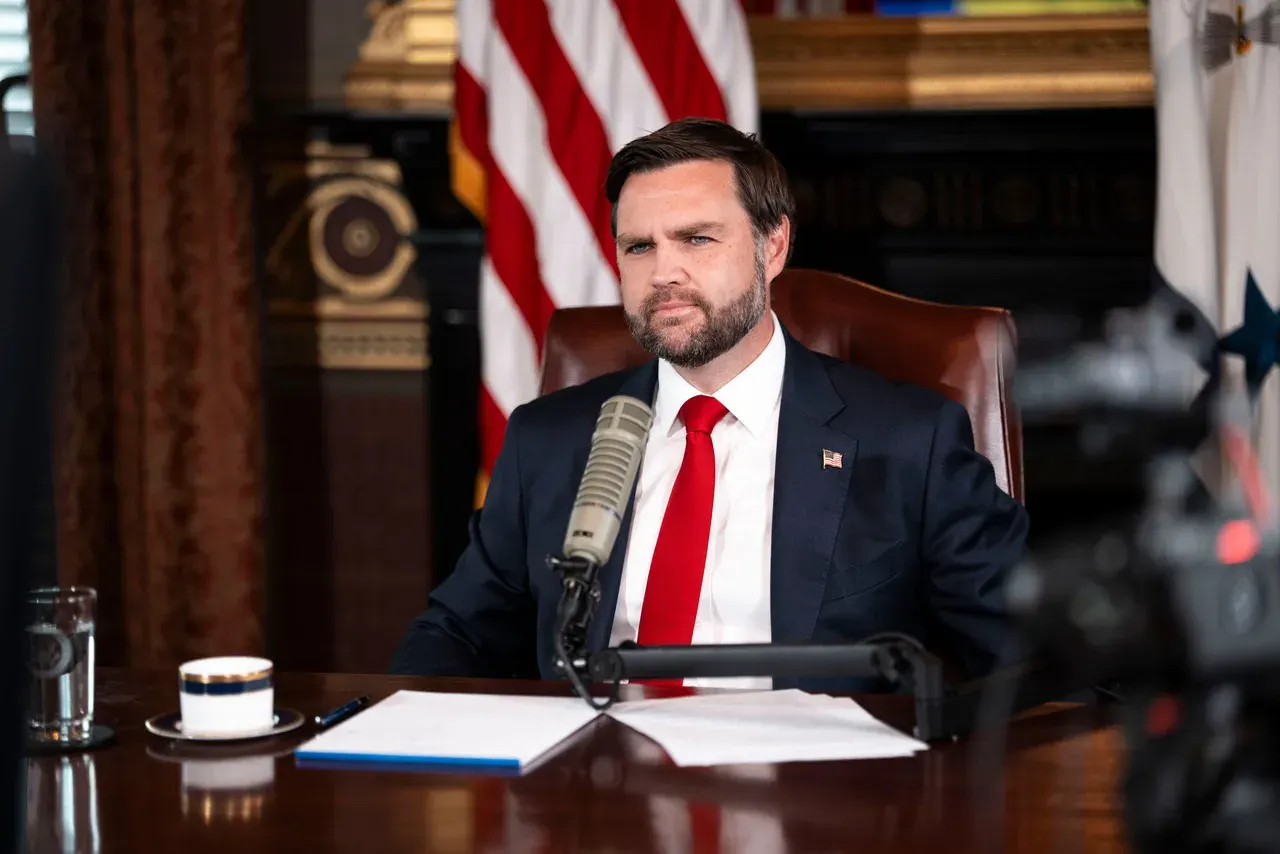 Bearded man in a dark suit and red tie seated at a microphone in an office, with American flags in the background