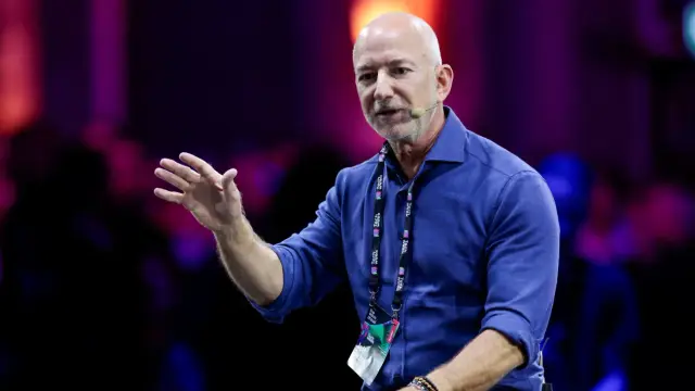 Bald man in a blue shirt and a lanyard speaks into a headset microphone with one arm extended, against a dark background with lights