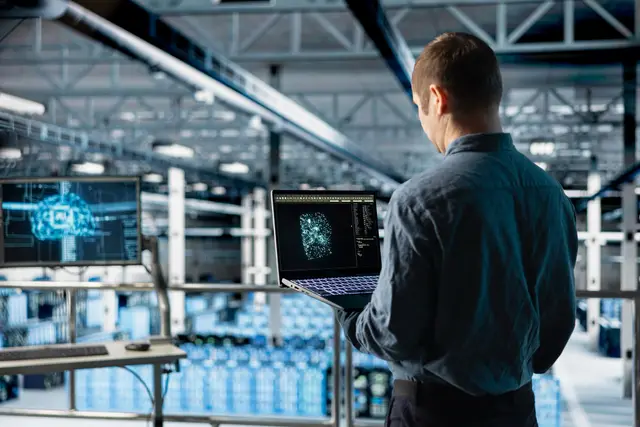 Man seen from behind holding an open laptop in an industrial hall, next to a screen showing a blue digital figure